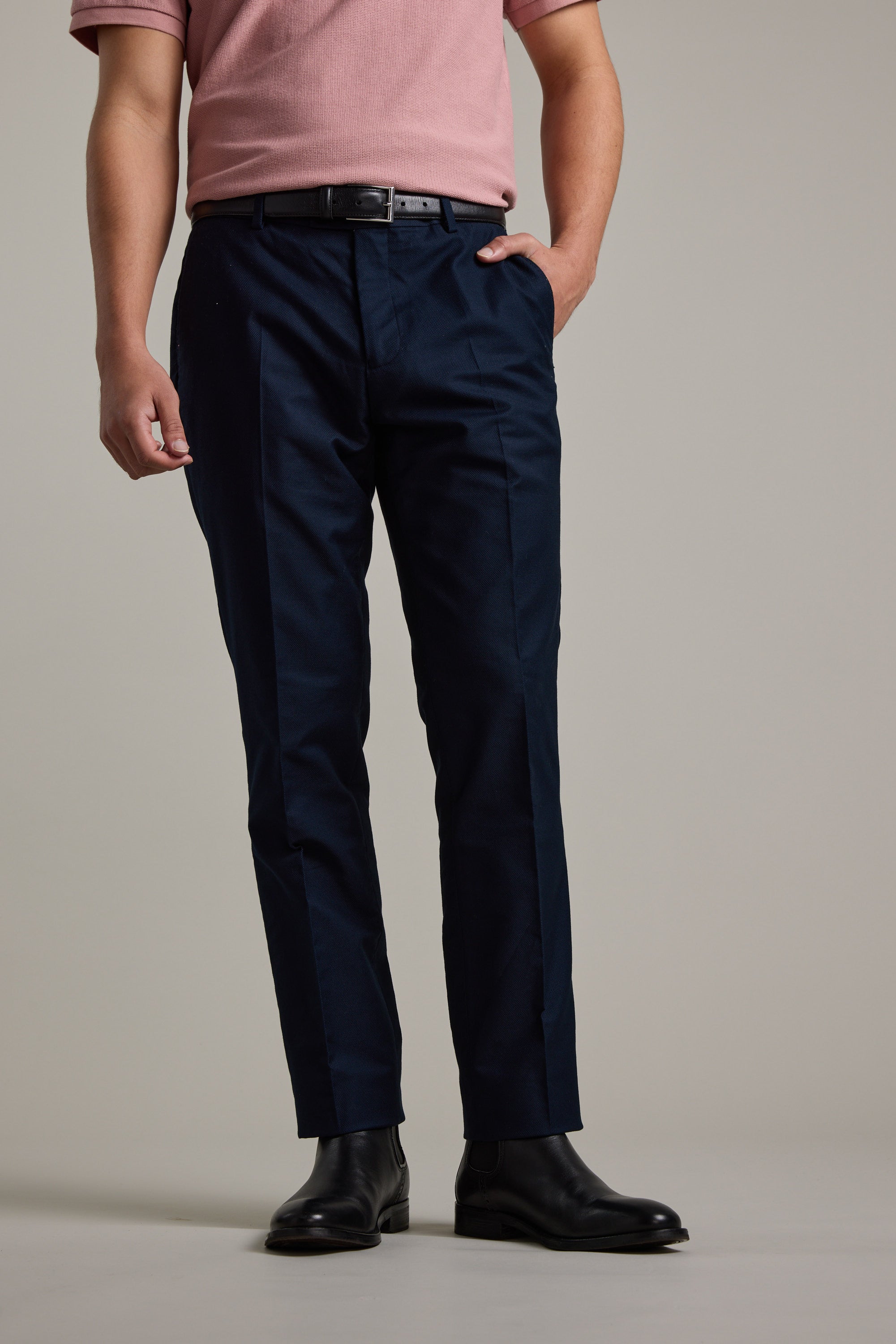 Wearing a tailored pink short-sleeve shirt, Moris Cotton Pant by Barkers in dark blue, a black belt, and black dress shoes, a person stands with one hand in their pocket against a plain background.