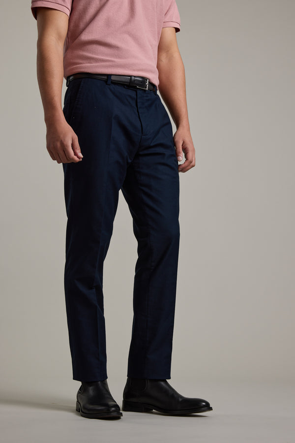 A man stands against a plain background wearing a light pink tailored shirt, black shoes, and Barkers Moris Cotton Pant in navy blue with a belt. His face is not shown.