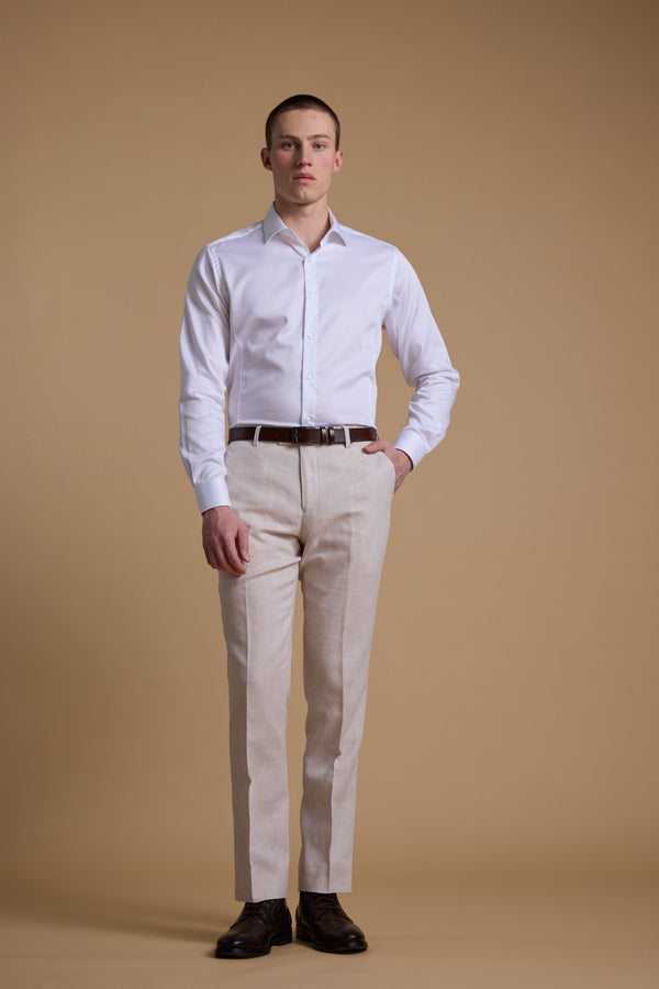 A man stands against a beige background wearing Barkers' Somerton Texture Pant, a light beige summer trouser in a breathable linen-cotton blend, paired with a white shirt, brown belt, and dark brown dress shoes, looking straight ahead.