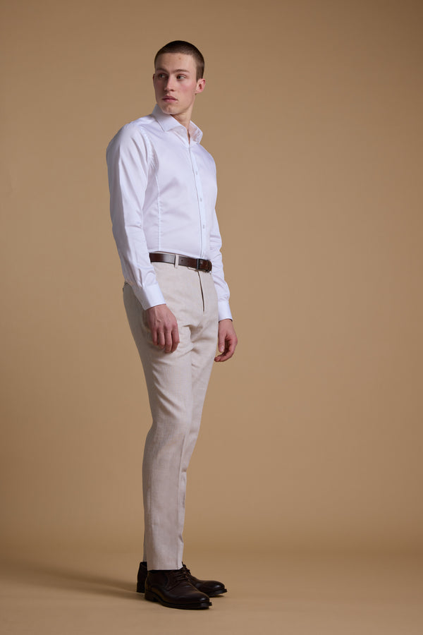 A man stands against a plain beige background wearing Barkers' Somerton Texture Pant in a breathable linen-cotton blend, paired with a white dress shirt, brown belt, and dark brown dress shoes, looking slightly to his left.