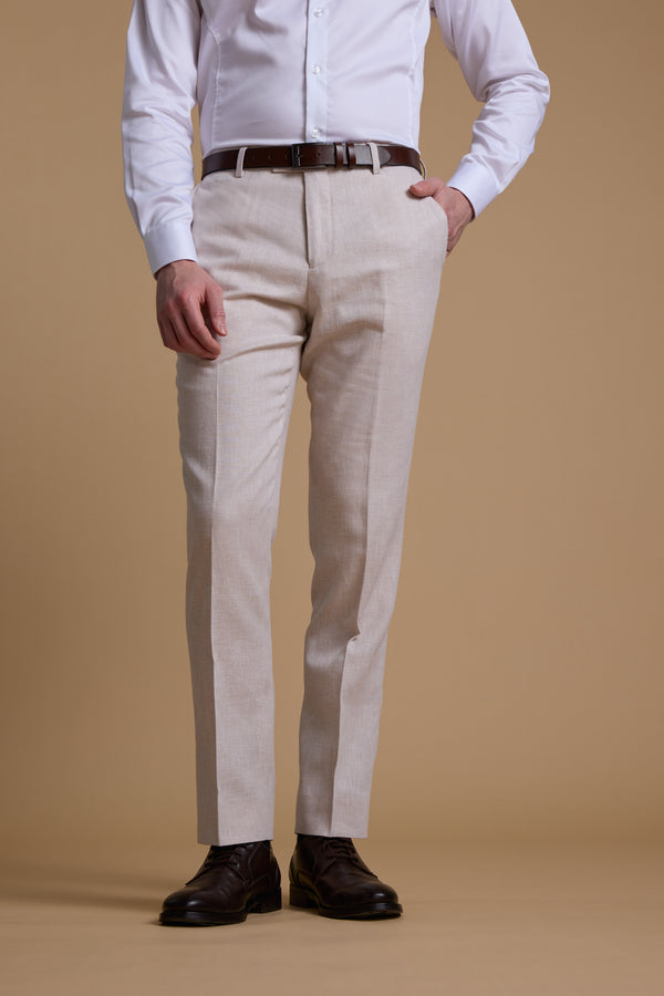 A man models the Barkers Somerton Texture Pant in beige, paired with a white shirt, brown belt, and brown dress shoes. He stands against a tan background with one hand in his pocket; his face is not shown.