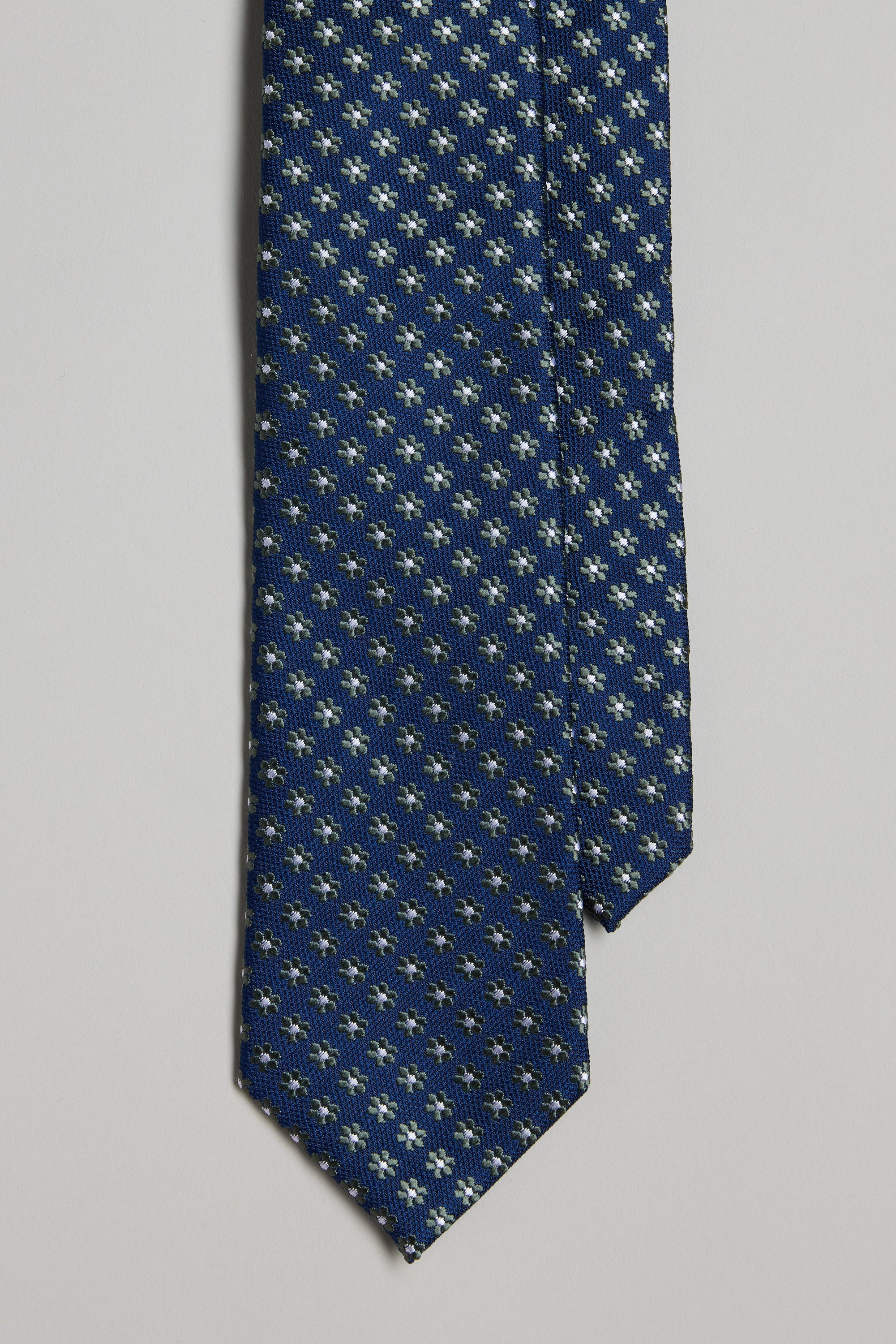 A close-up of the Barkers Carmen Flower Tie in navy, showcasing a repeating small white and light yellow pattern—ideal with silk accessories such as pocket squares or bow ties—displayed against a plain light background.