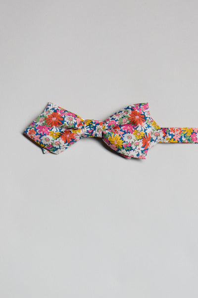 The Liberty Libby Bow Tie by Liberty features a vibrant floral print in red, orange, pink, yellow, and white on a light background. Handmade in New Zealand and shown on a plain white surface.