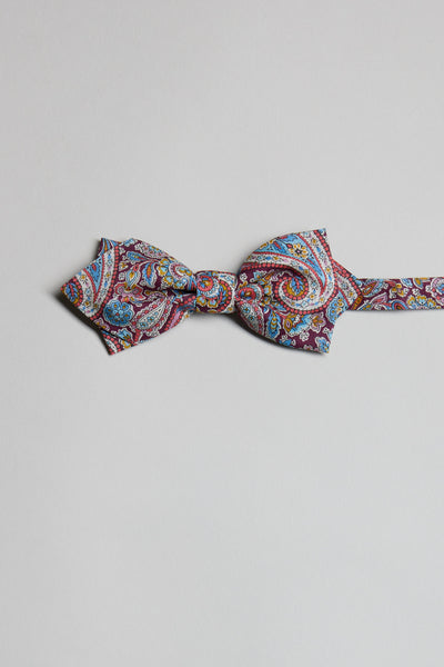 The Liberty Lee Manor Bow Tie by Liberty features a vibrant paisley print in red, blue, yellow, and white tones. Handmade in New Zealand, it is showcased against a plain light gray background.