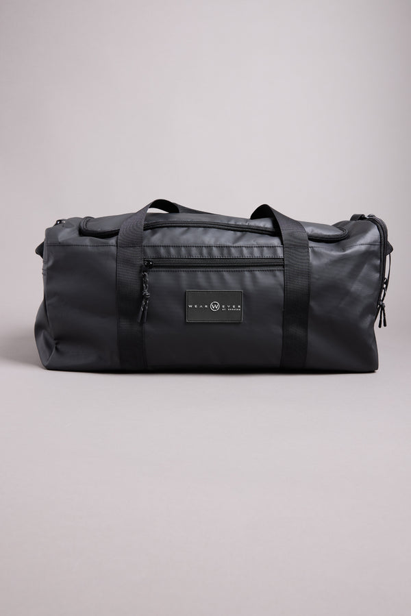 The WearEver Transit Holdall, a black water-resistant duffel bag with black handles, a front zipper pocket, and a rectangular logo patch, is shown against a plain light gray background.