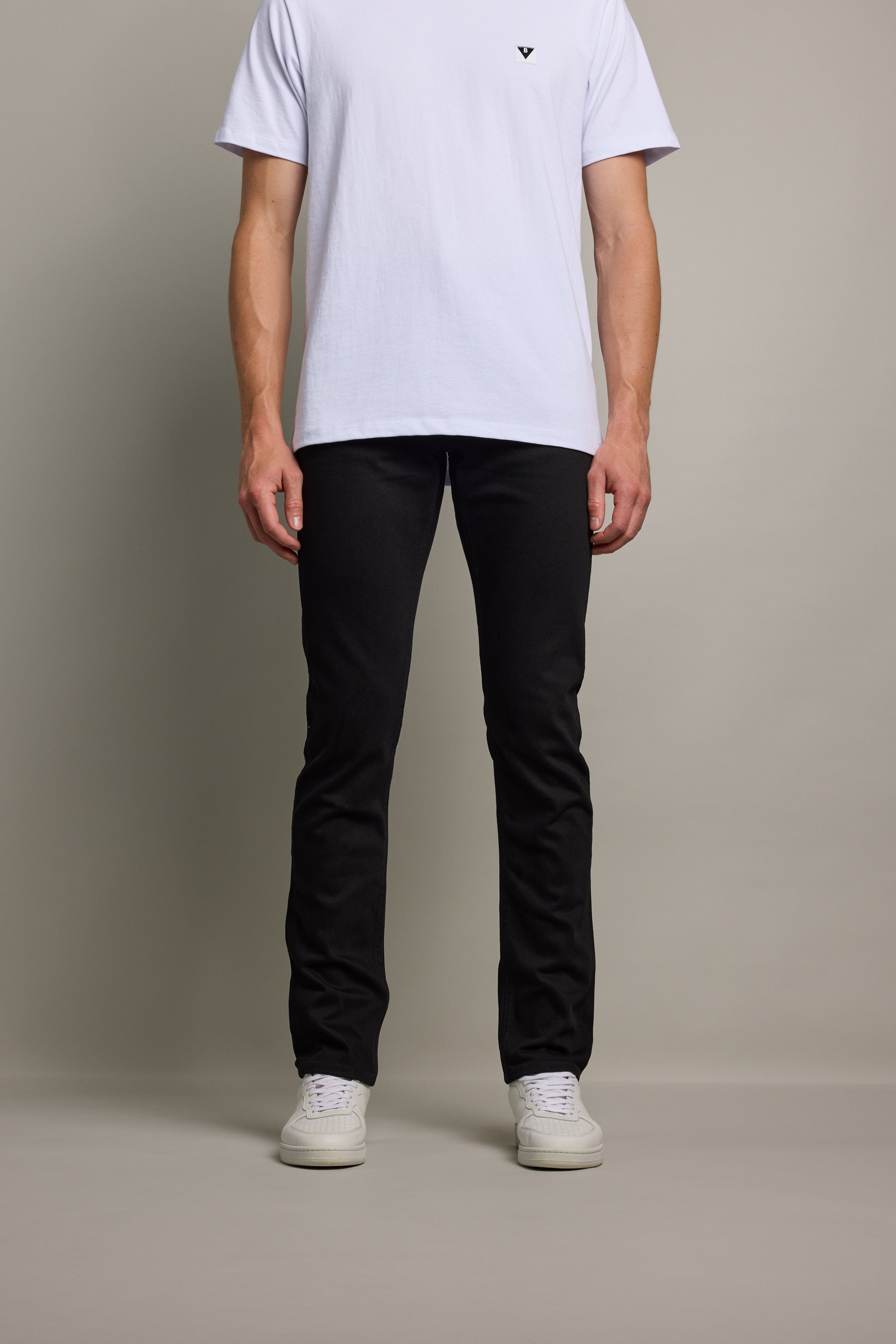 A person stands facing forward against a plain background, wearing a white short-sleeve T-shirt, Barkers Rambler Denim Slim Jean, and white sneakers. Only the body from shoulders down is visible.