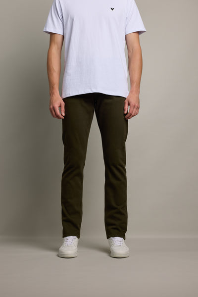 A person, head not visible, stands against a plain background wearing a white t-shirt, Barkers Nomad Taper Coloured Jean in dark olive green, and white sneakers.