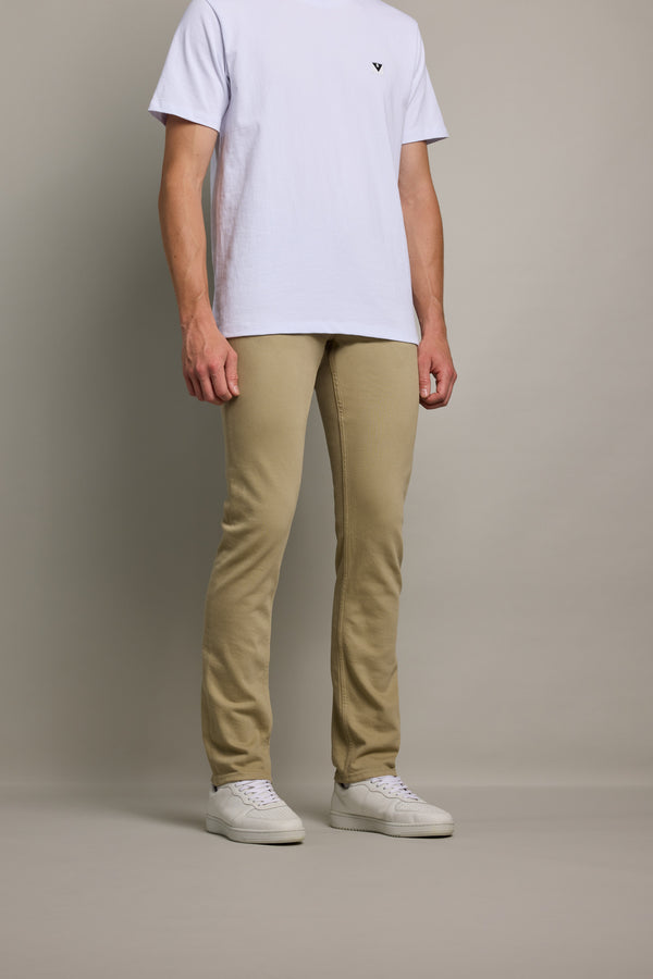 A person wearing Barkers' Rambler Slim Coloured Jean in tan, a plain white T-shirt, and white sneakers stands against a neutral background. The image is cropped from the shoulders down.