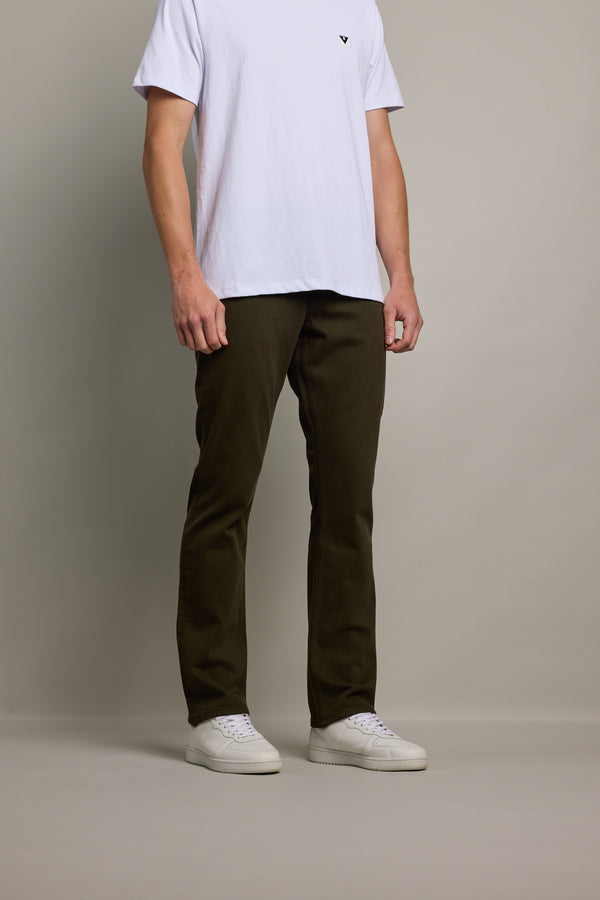 A person stands against a plain background wearing a white short-sleeve t-shirt, Barkers Wanderer Straight Coloured Jean in dark olive made from regenerative cotton, and white sneakers. The person’s face is not visible.