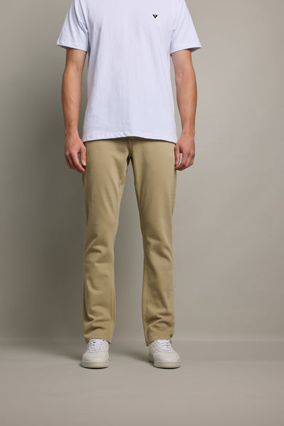 A person stands against a plain background, wearing a white T-shirt, Barkers Wanderer Straight Coloured Jeans made from regenerative cotton in beige, and white sneakers. The photo is cropped from shoulders down.
