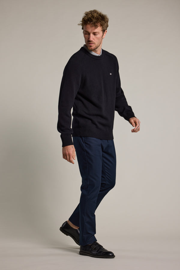 A man with short, tousled hair wears a black sweater, Barkers’ Lyndon Cotton Pants in blue, and black shoes. He stands against a plain light backdrop, looking down to his right for a smart casual look.