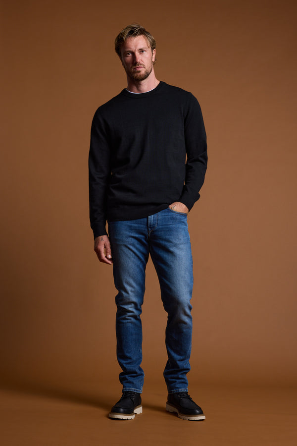 Man in Barkers Skyland ZQrx Merino Crew and blue jeans stands against a brown background.