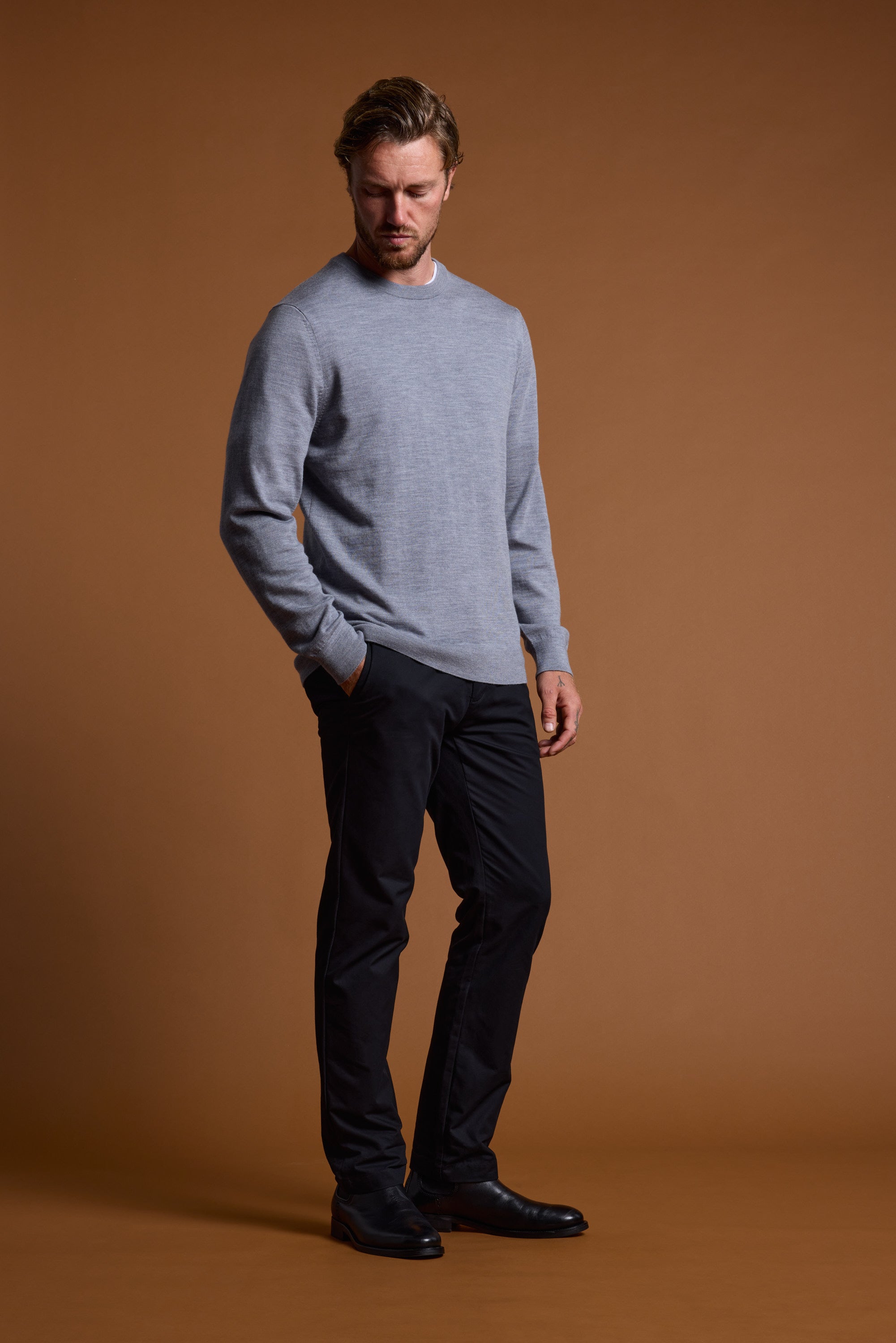 A man stands against a brown background, looking down with one hand in his pocket. He is wearing a light gray Barkers Skyland ZQrx Merino Crew made from ethically sourced New Zealand merino wool, paired with dark pants and black shoes.