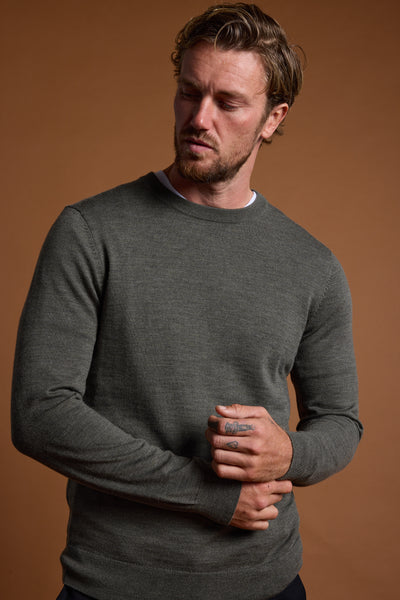 A man with wavy brown hair and a short beard wears the Barkers Skyland ZQrx Merino Crew, made from ethically sourced New Zealand merino wool, standing against a brown background and looking down to his left while holding one hand with the other.