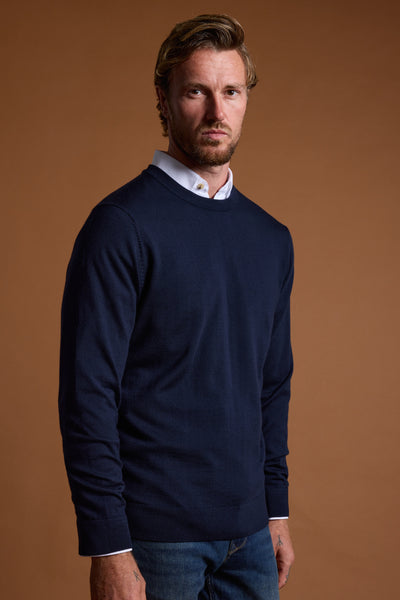 A man with light brown hair and a beard wears a white collared shirt under a Barkers Skyland ZQrx Merino Crew, made from sustainable New Zealand merino wool, and blue jeans, standing against a plain brown background while looking at the camera.