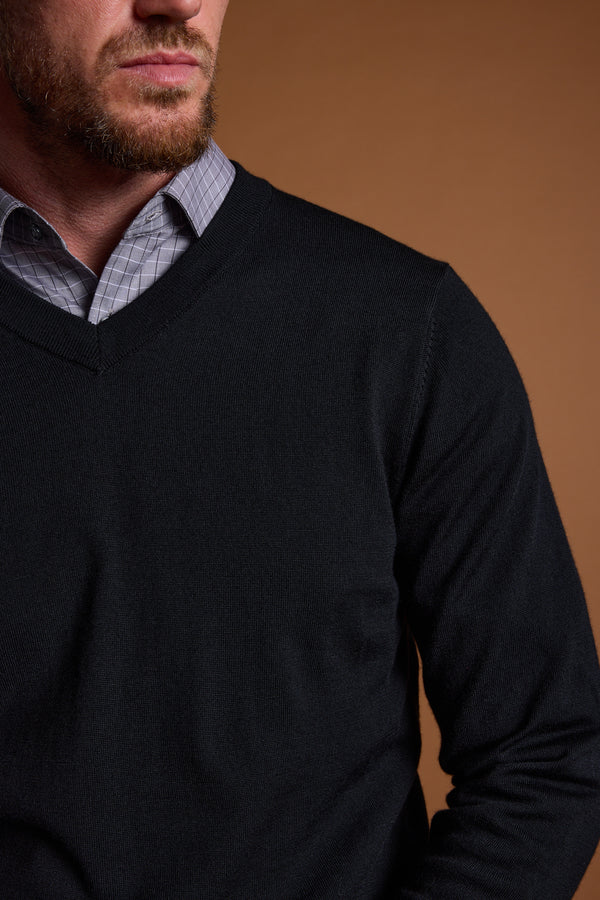 A man in the Barkers Skyland ZQrx Merino Vee, a dark V-neck sweater made from traceable merino, layered over a checkered collared shirt, is shown from the shoulders up with his face partly visible against a brown background.