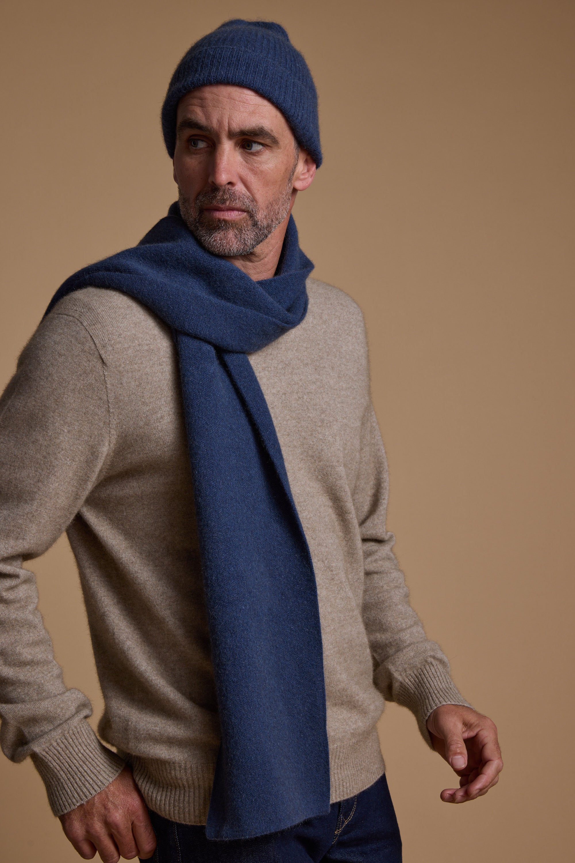 A man in a beige sweater accessorizes with the NZ Made Perino Beanie and matching scarf by Woolyarns Perino, both in a blue merino possum silk blend, standing against a tan background and looking to the side with a neutral expression.