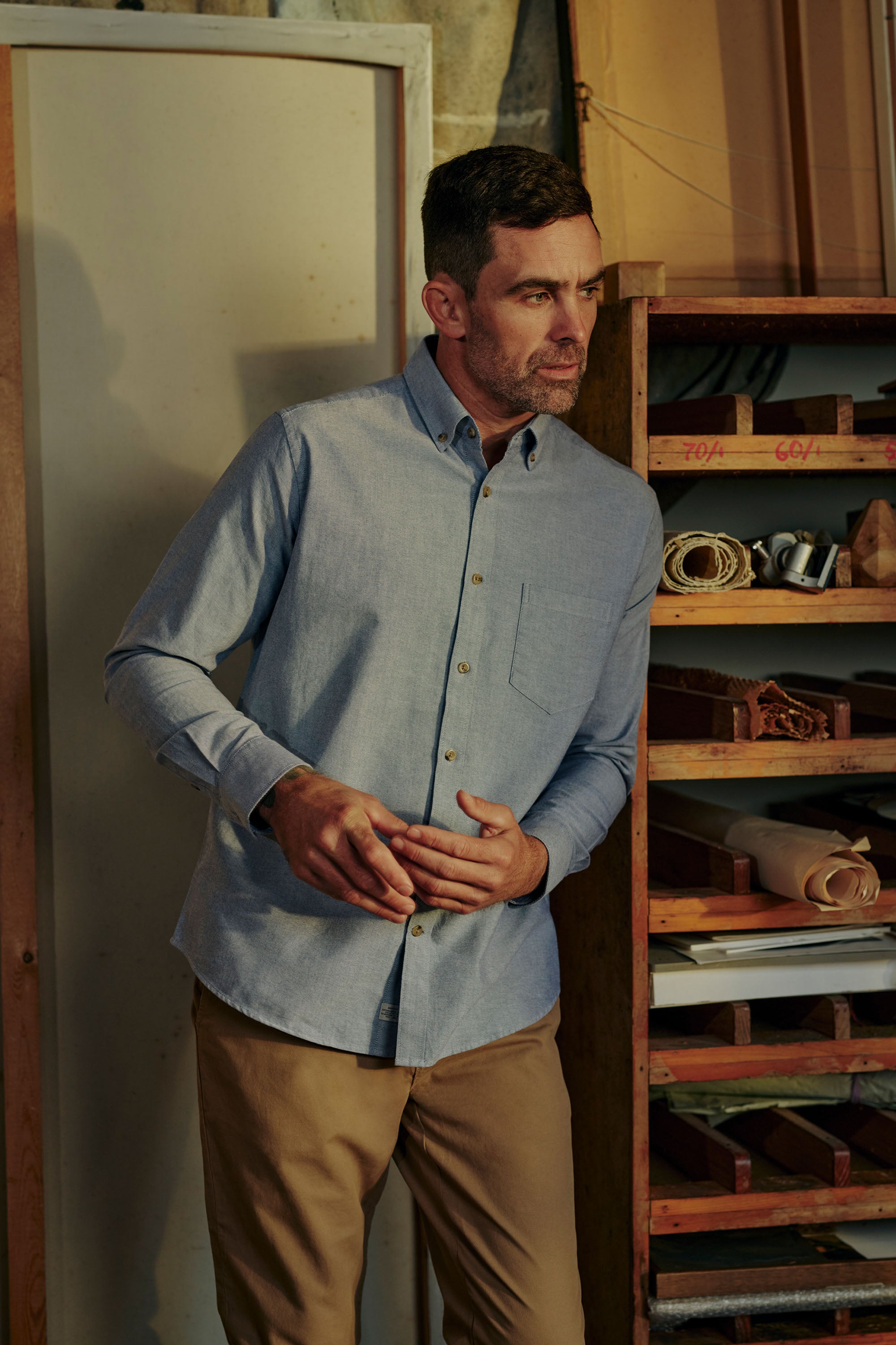 A man in the Barkers Thatcher Oxford Shirt, crafted from responsibly sourced cotton, and khaki pants stands indoors by wooden shelves lined with tools and materials, gazing thoughtfully to the side.