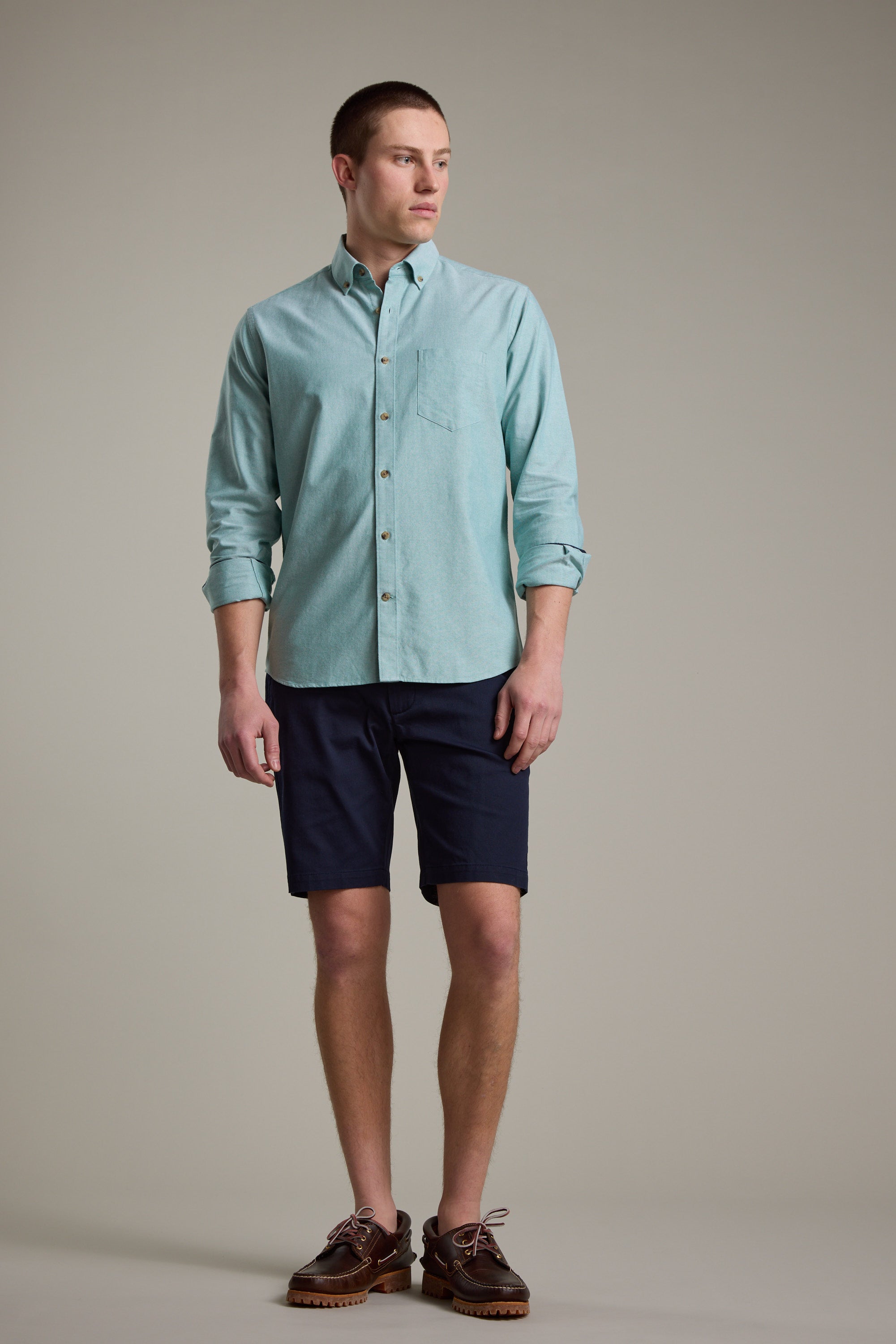A young man stands against a plain background, wearing the Barkers Thatcher Oxford Shirt in light blue with rolled-up sleeves, paired with dark navy shorts and brown boat shoes. He looks to his left with a neutral expression.