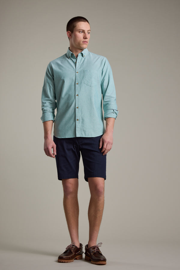 A young man stands against a plain background, wearing the Barkers Thatcher Oxford Shirt in light blue with rolled-up sleeves, paired with dark navy shorts and brown boat shoes. He looks to his left with a neutral expression.