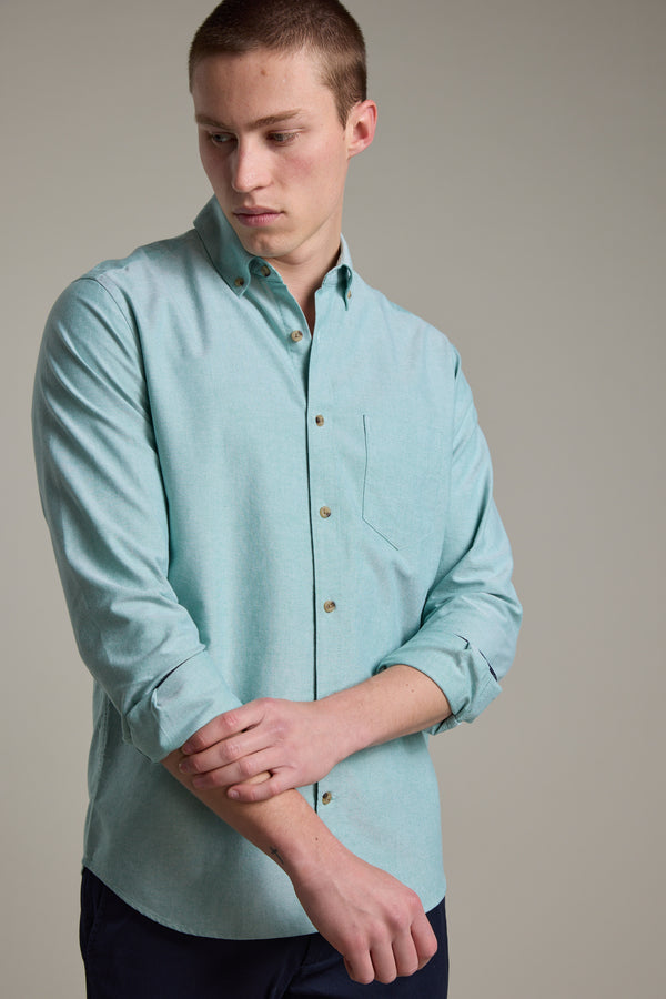 A young man in a Barkers Thatcher Oxford Shirt, tailored fit and light teal with rolled-up sleeves, adjusts his left sleeve against a plain light-grey background.