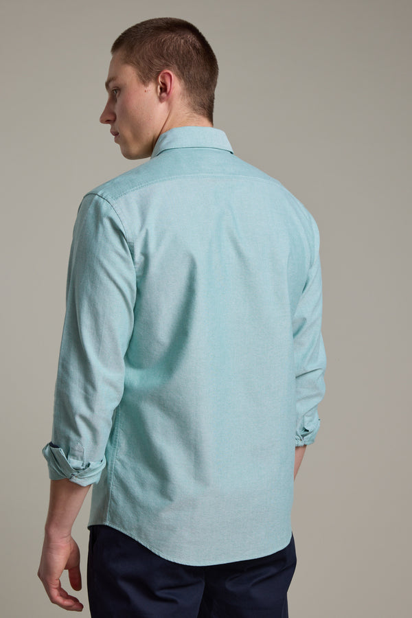 A person with short brown hair wears the Barkers Thatcher Oxford Shirt in light blue, made from responsibly sourced cotton, paired with dark pants, standing facing away from the camera against a plain background.