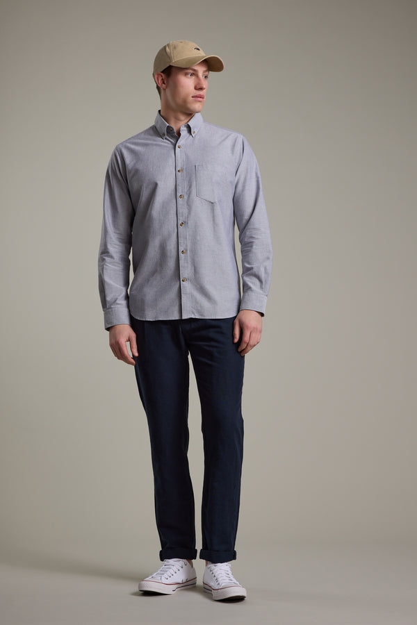 A man stands against a plain background wearing a beige baseball cap, dark blue pants, white sneakers, and the Barkers Thatcher Oxford Shirt in light gray tailored fit made from responsibly sourced cotton. He looks slightly to the side with relaxed arms.