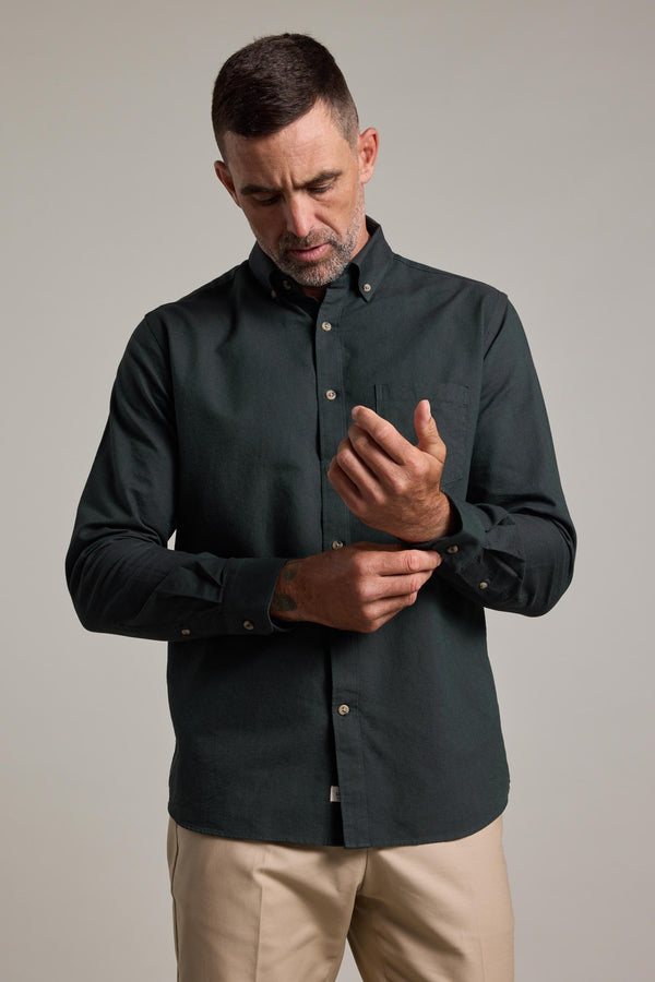A man with short dark hair and a trimmed beard wears the Barkers Thatcher Oxford Shirt in dark green with beige pants, looking down as he adjusts his shirt cuff against a plain light background.