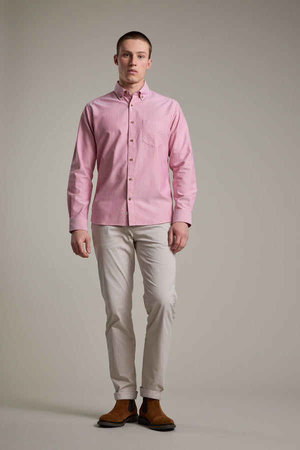 A man with short hair and a neutral expression stands against a plain background wearing the Barkers Thatcher Oxford Shirt in light pink (100% cotton), paired with light beige tailored fit pants and brown boots.