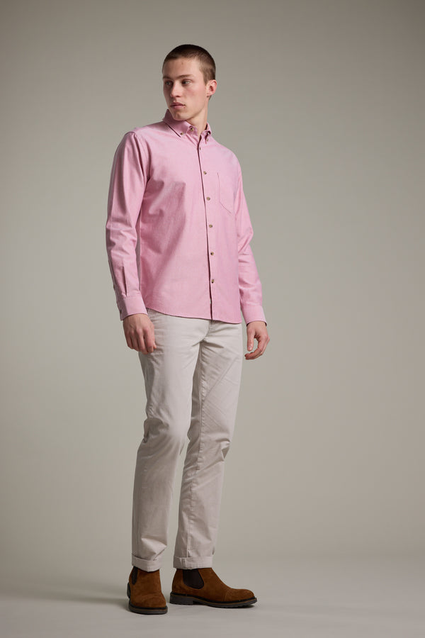 A man stands slightly turned, wearing the Barkers Thatcher Oxford Shirt in light pink, paired with light beige pants and brown suede shoes against a plain gray background.