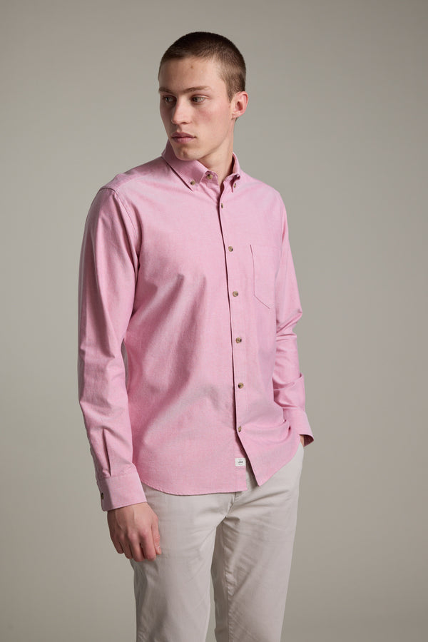 A young man wears the Barkers Thatcher Oxford Shirt in light pink, tailored fit, made from responsibly sourced cotton with a chest pocket, paired with light beige pants. He stands against a plain gray background, looking slightly left with a neutral expression.