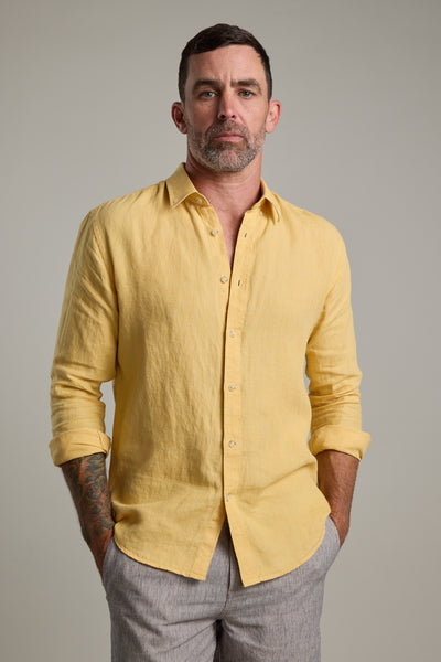 A man with short dark hair, a trimmed beard, and tattoos on his left forearm wears the Barkers Dorset Linen Shirt in yellow and light gray pants, standing with hands in his pockets against a plain background.