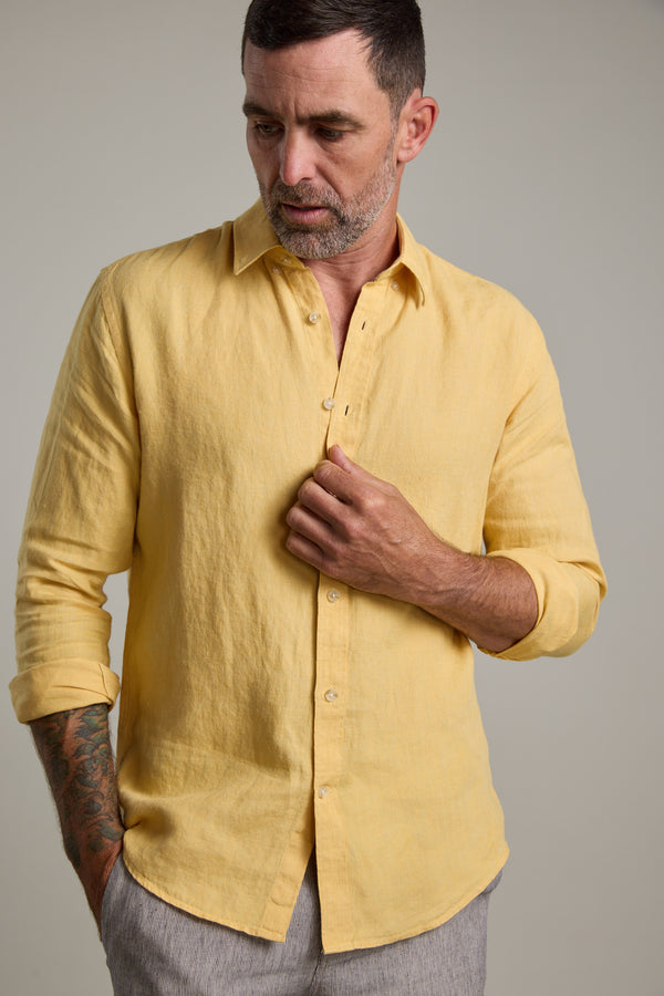 A man with short dark hair and a beard wears the Barkers Dorset Linen Shirt in light yellow and light gray pants. He looks down while fastening a button on his shirt.