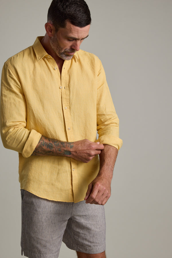 A man with short dark hair and a beard wears the Barkers Dorset Linen Shirt in yellow and gray shorts. He rolls up his sleeve, revealing a tattoo on his left forearm, while standing against a plain, light-colored background.