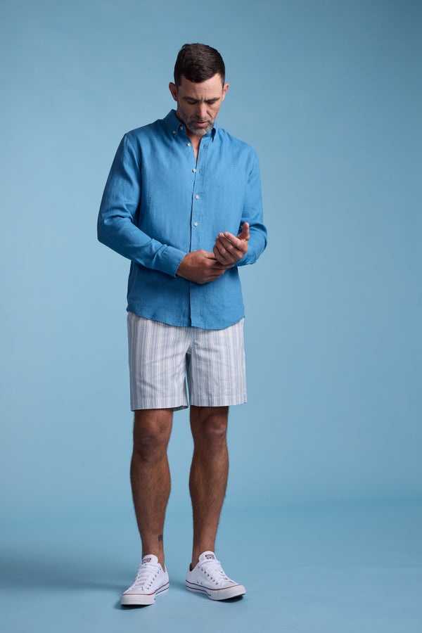 A man in a Barkers Dorset Linen Shirt, paired with striped shorts and white sneakers, stands against a plain blue background, looking down and holding his left wrist with his right hand.