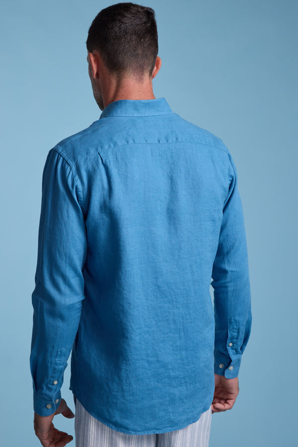 A man stands with his back to the camera in a bright blue Barkers Dorset Linen Shirt and light-striped pants, set against a plain light blue background.