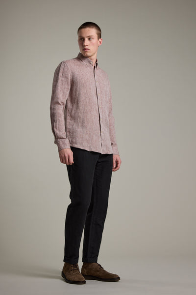 A young man stands against a plain gray background, wearing the Barkers Dorset Linen Shirt in light pink with a tailored fit, paired with black pants and brown suede shoes, looking slightly to his right with a neutral expression.