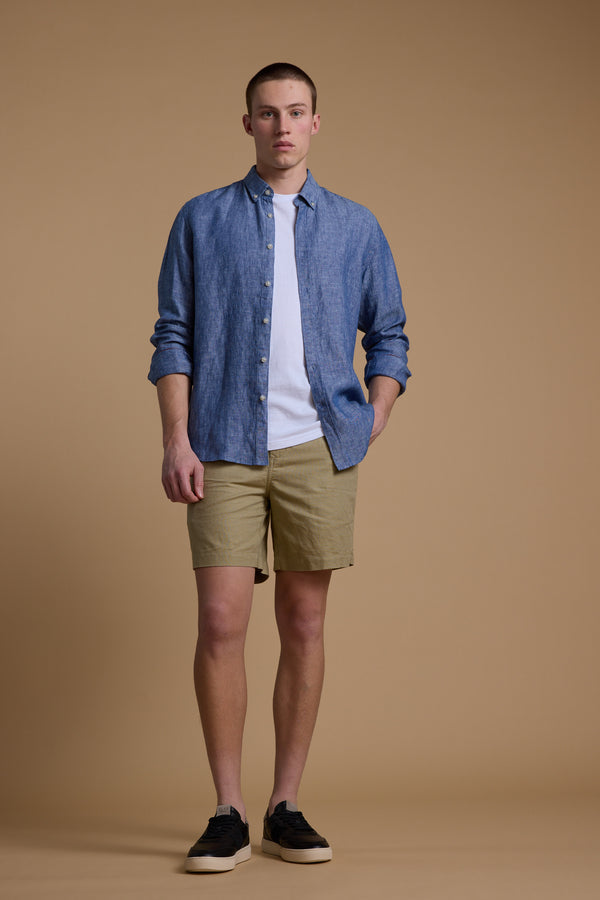 A young man with short hair and a neutral expression stands against a beige background wearing the Barkers Dorset Linen Shirt in blue over a white tee, tan shorts, and black sneakers, with one hand in his pocket.