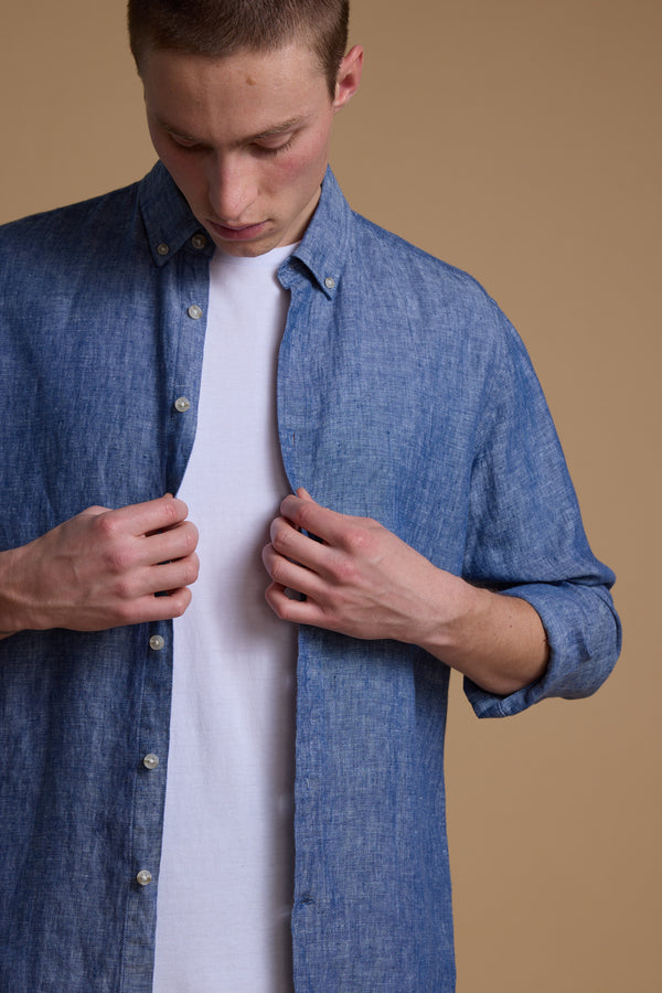 A person wearing the Barkers Dorset Linen Shirt in blue over a white tee buttons it up, showcasing its tailored fit and breathable linen against a solid beige background.