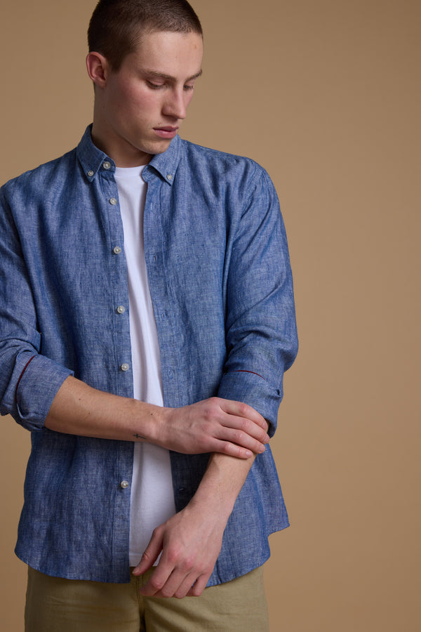 A young man stands against a beige background, looking down as he rolls up the sleeve of his Barkers Dorset Linen Shirt in blue over a white tee, paired with light khaki pants.