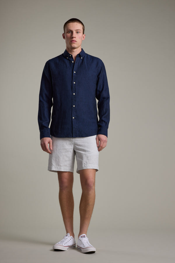 A man stands facing forward, wearing the Barkers Dorset Linen Shirt in dark blue, paired with light gray shorts and white sneakers against a plain, light-colored background.