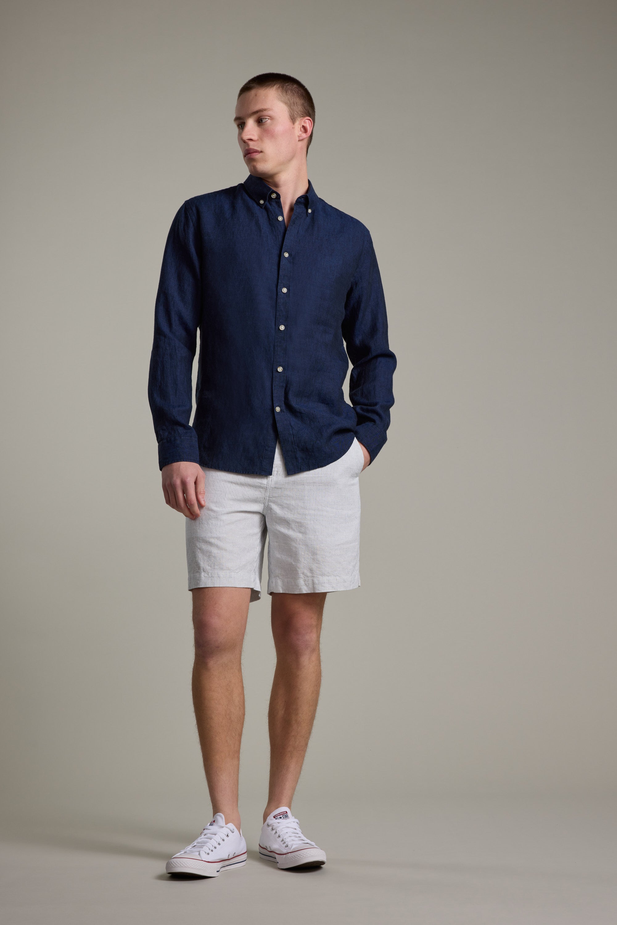 A man stands against a plain background wearing the Barkers Dorset Linen Shirt, tailored fit in dark blue, paired with light gray shorts and white sneakers. He has one hand in his pocket and looks slightly to the side.