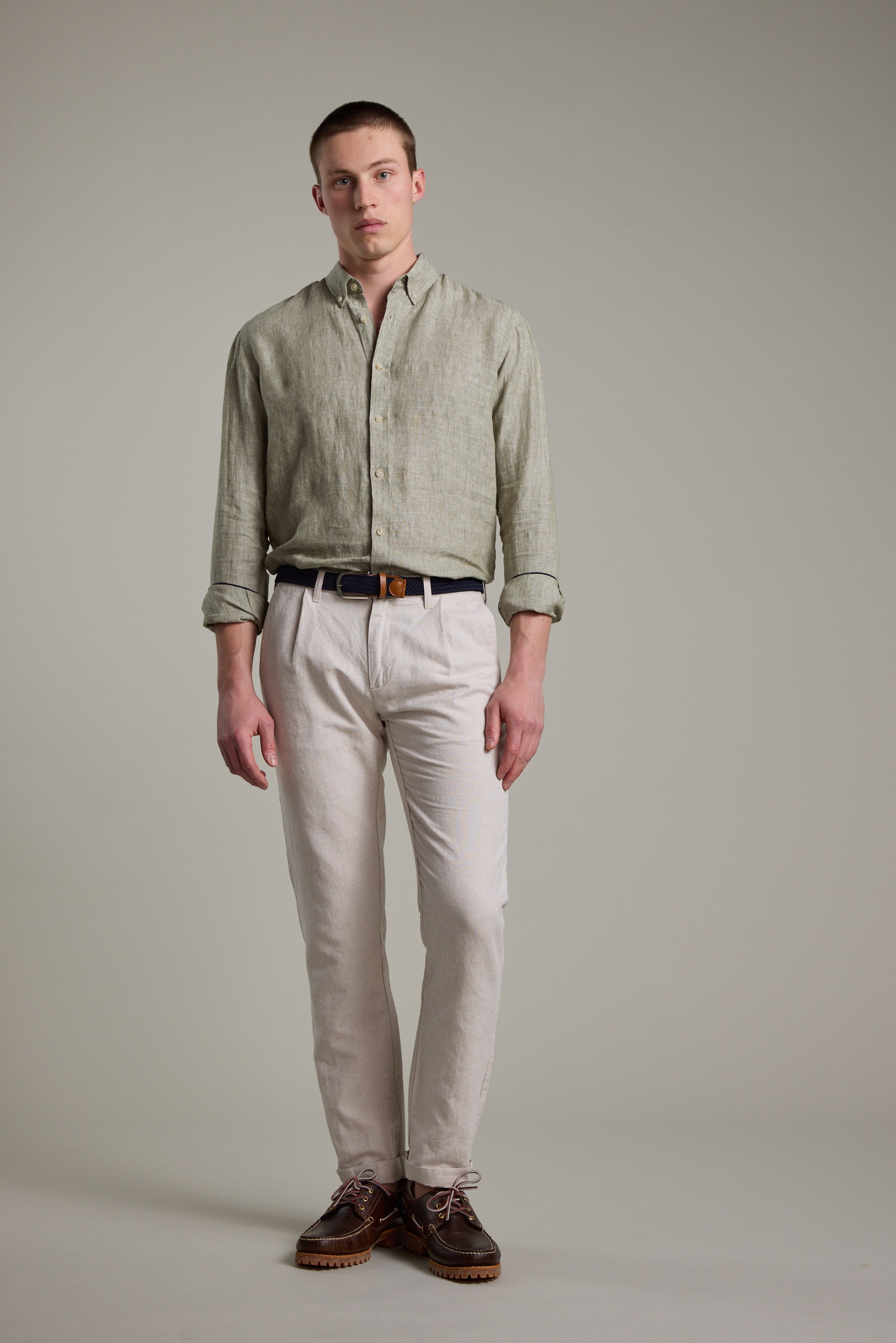 A young man with short hair stands against a plain background, wearing the Barkers Dorset Linen Shirt in light green with rolled sleeves, beige pants, a black belt, and brown shoes. The breathable fabric and tailored fit enhance his refined look.