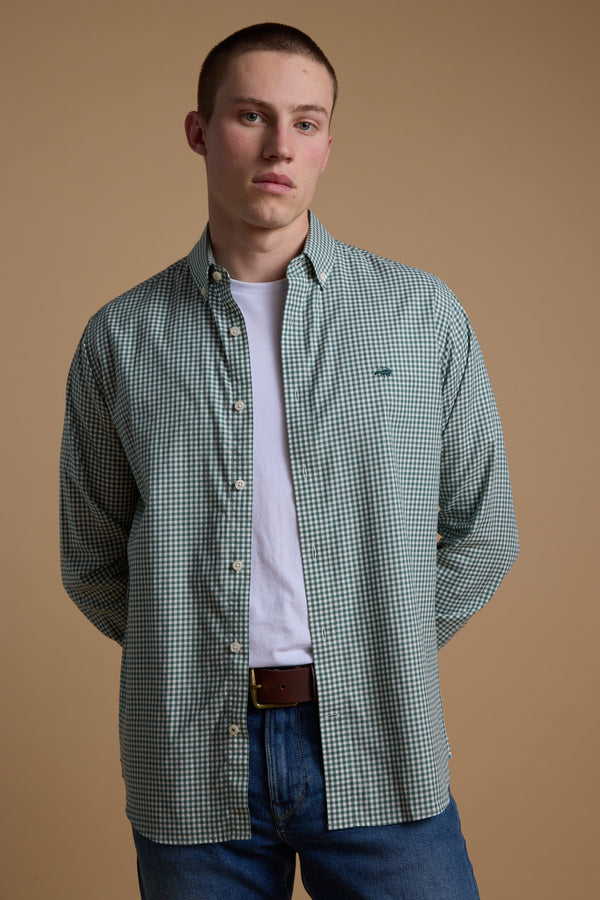 A young man with short hair stands against a beige background, wearing a white t-shirt, blue jeans, and the Barkers Courtland Check Shirt in green and white checks made from responsibly sourced cotton, with his hands behind his back.