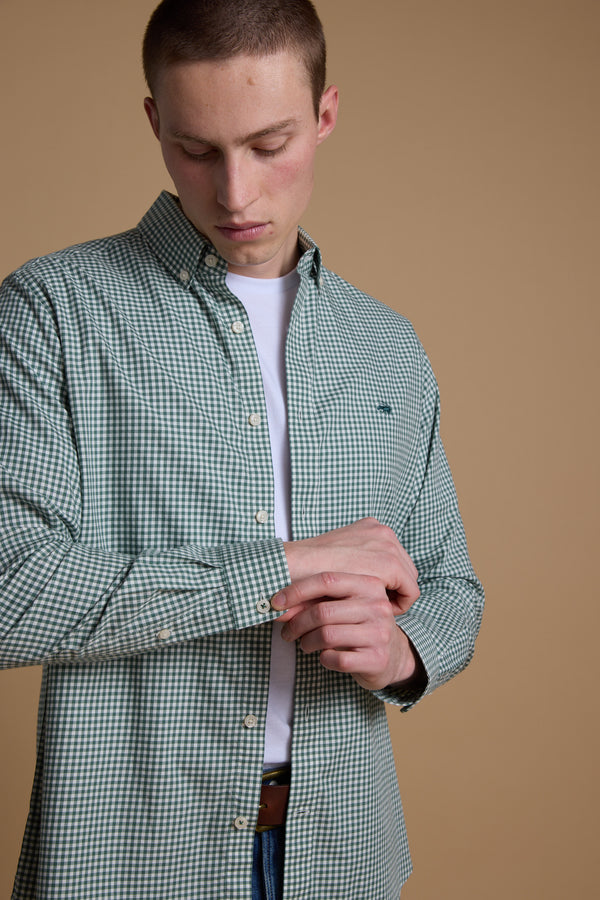 A man wearing a Barkers Courtland Check Shirt in green and white over a white T-shirt adjusts his shirt cuff while looking down against a plain beige background.