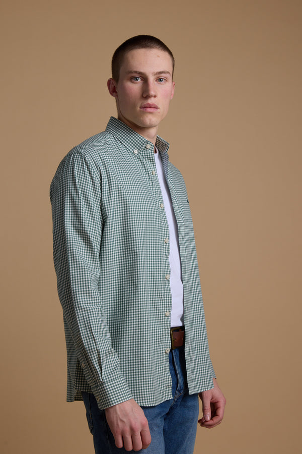 A young man with short hair wears a green checkered Barkers Courtland Check Shirt over a white t-shirt and blue jeans, standing against a plain beige background and looking directly at the camera.