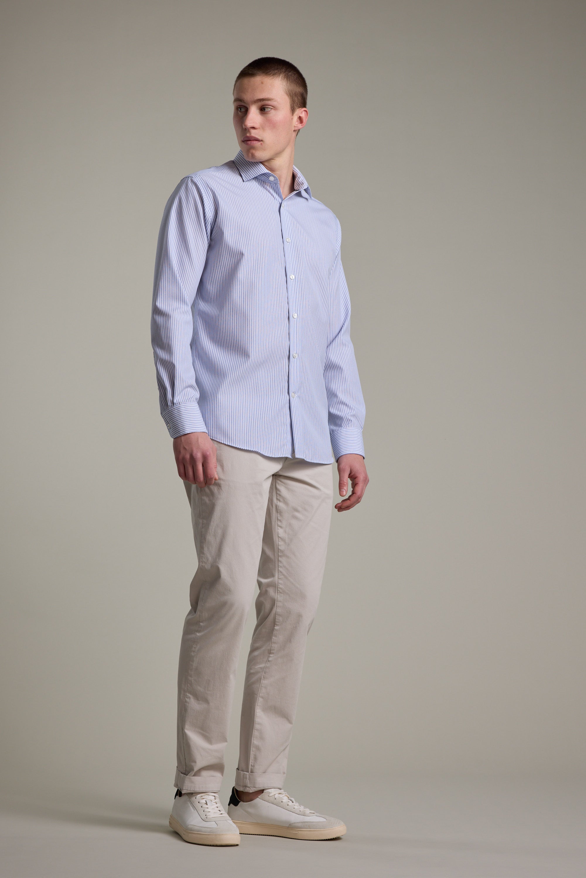A young man stands against a plain background wearing the Barkers Norwoord Stripe Business Shirt, beige chinos, and white sneakers. He looks slightly to his left with a neutral expression.