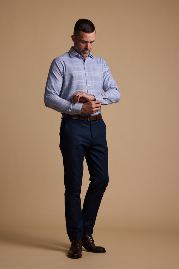A man stands against a beige background, wearing the Barkers Kenmore Check Business Shirt in light blue with dark trousers and brown shoes, glancing down at his watch with one arm raised.
