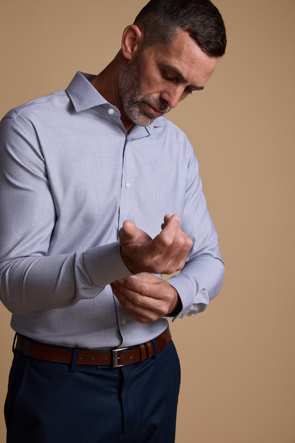 A man with short dark hair and a beard buttons the cuff of his light blue Barkers Eaton Jacquard Business Shirt, paired with dark pants and a brown belt, standing against a plain beige background.