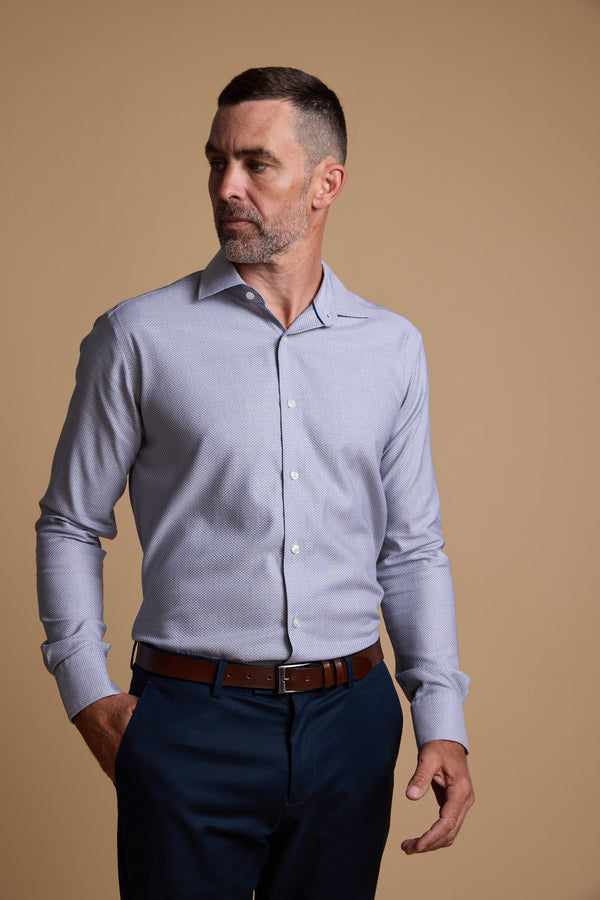 A man with short dark hair and a trimmed beard wears a light blue Barkers Eaton Jacquard Business Shirt, tailored fit trousers, and a brown belt. He stands against a beige background, looking left with one hand in his pocket.